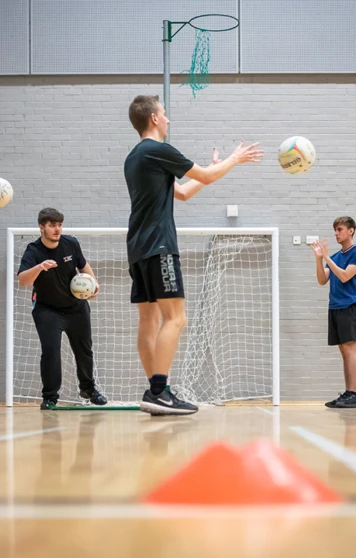 Students in a games hall tossing netballs in a circle as part of a group activity. Students in a games hall tossing netballs in a circle as part of a group activity.